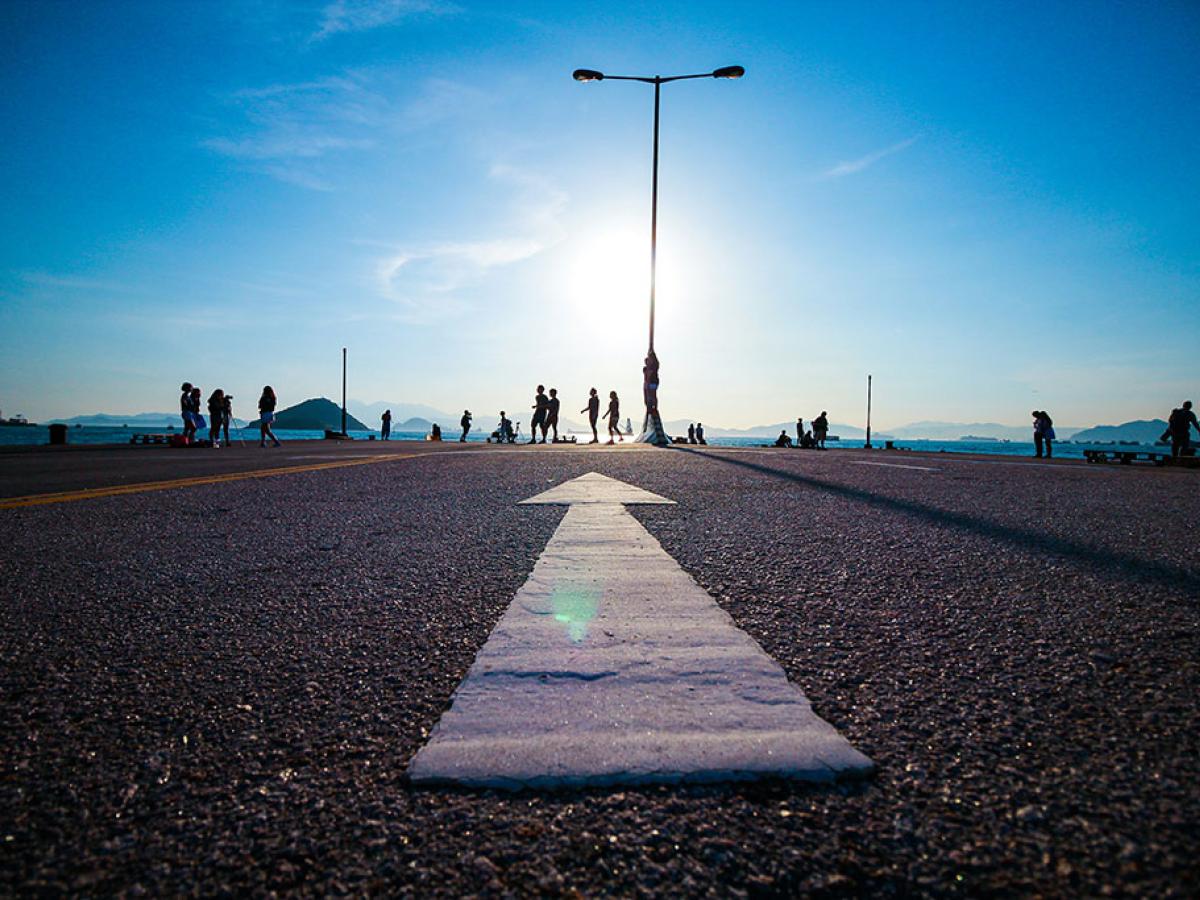 A white arrow on tarmac points towards a setting sun and people walking by a silhouetted lamp post.