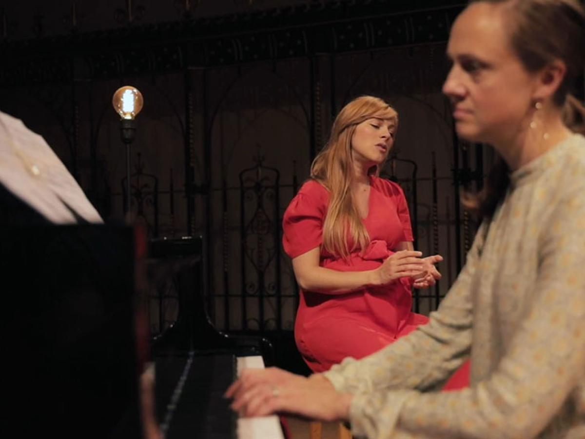 A pianist plays in the foreground and a seated singer gestures with eyes closed behind 