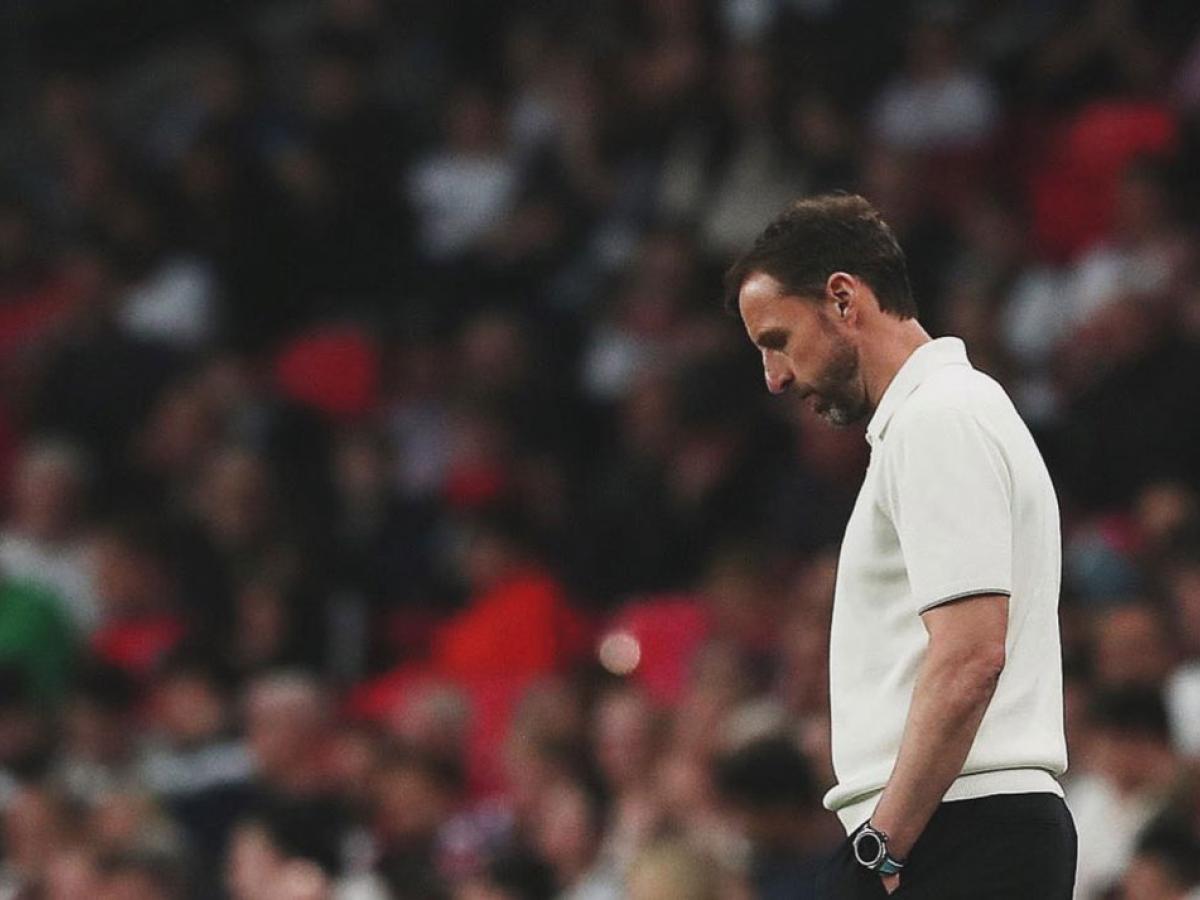 A dejected looking football manager ponders his feet while standing beside a pitch.