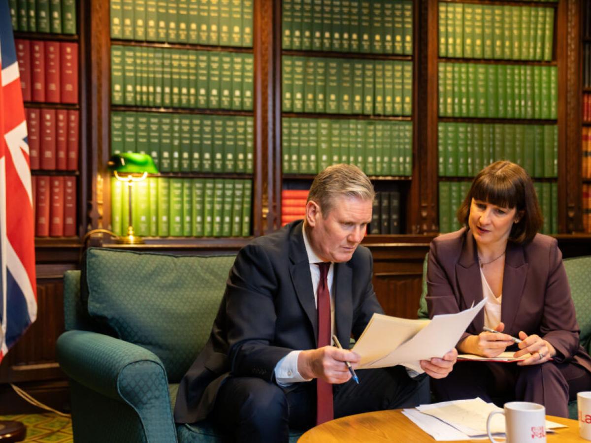 Kier Starmer sits on a sofa, leaning forward and holding papers he is reading. Rachel Reaves sits and looks on.