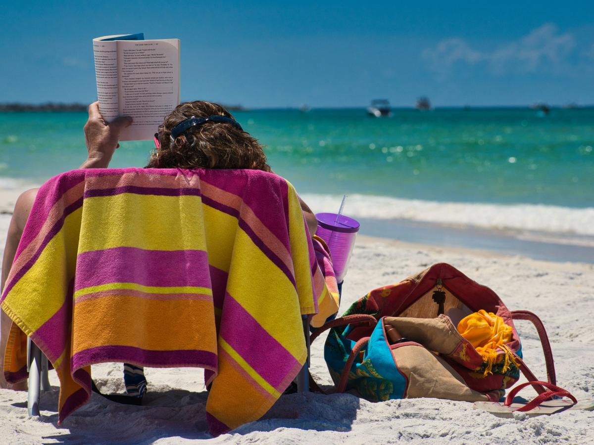 On a beach lounger someone holds a book aloft to read.