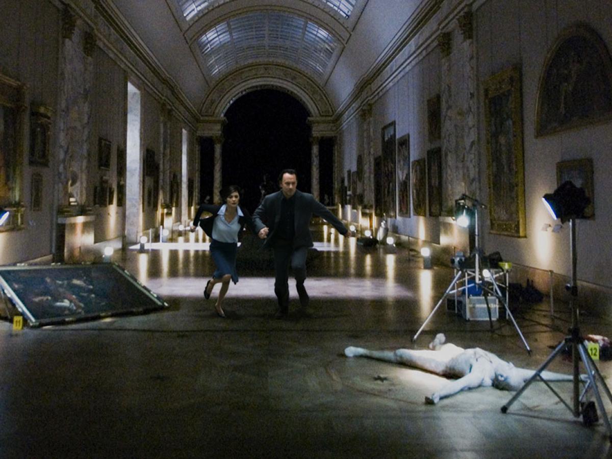 Two people run though a darkened art gallery towards a body lying amongst photography paraphanalia