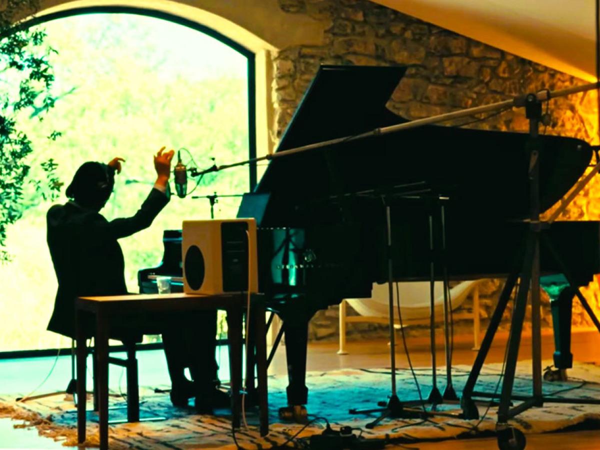 A pianist raises his arms while sitting at a grand piano amid recording equipment.