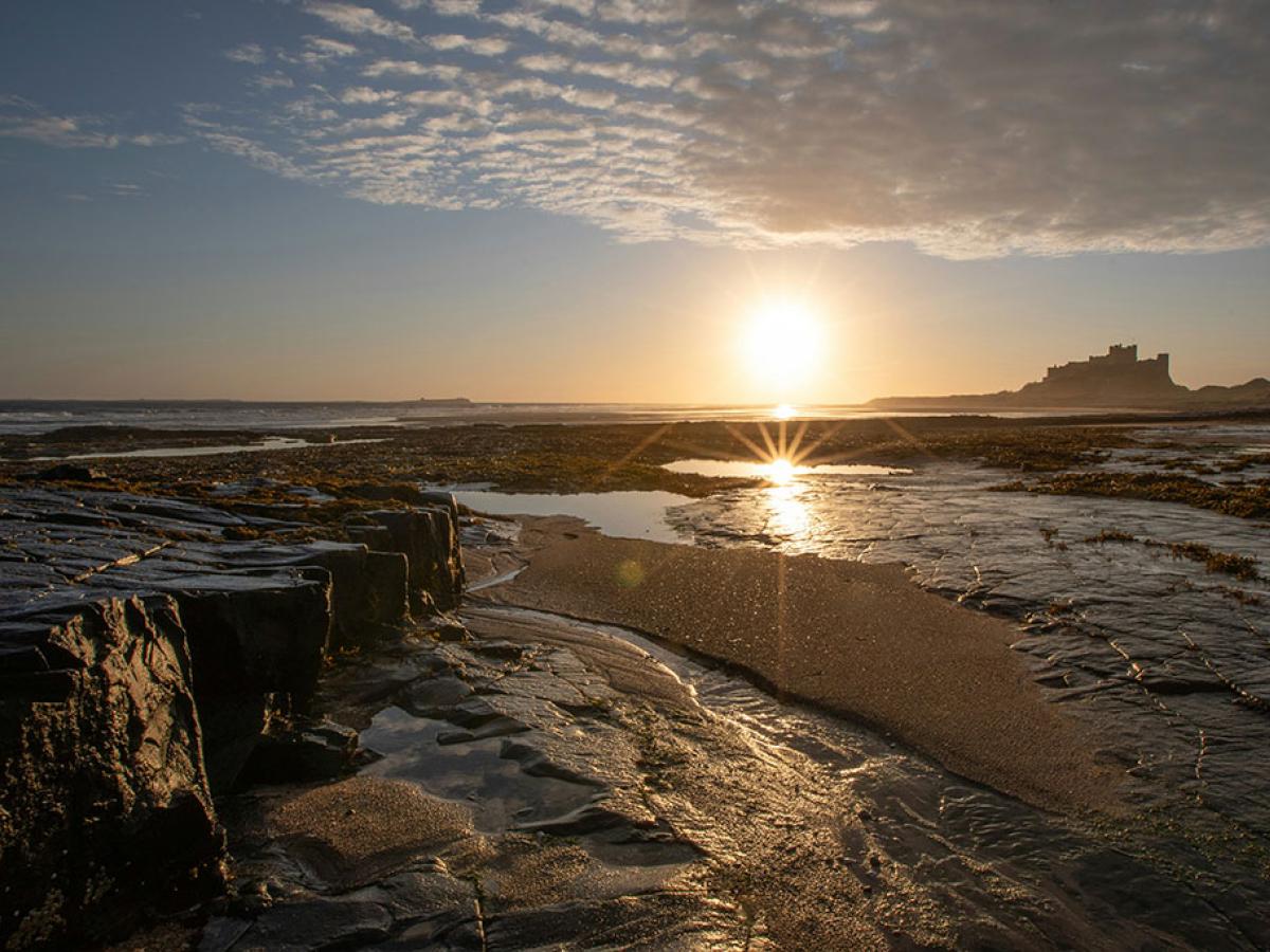 A rockpool on a beach reflects the sun, a castle stands beyond the sand dunes