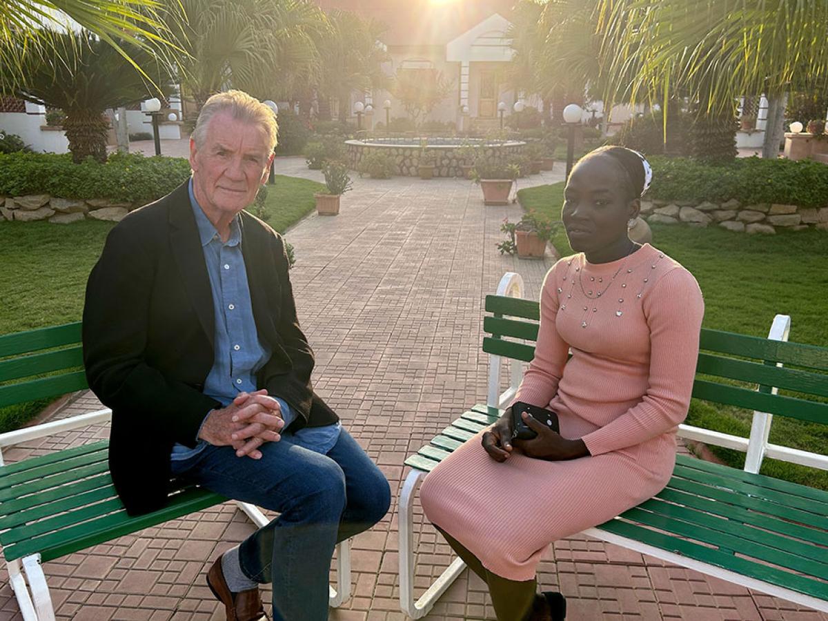 A TV interviewer sits across from a young woman in an outdoor setting