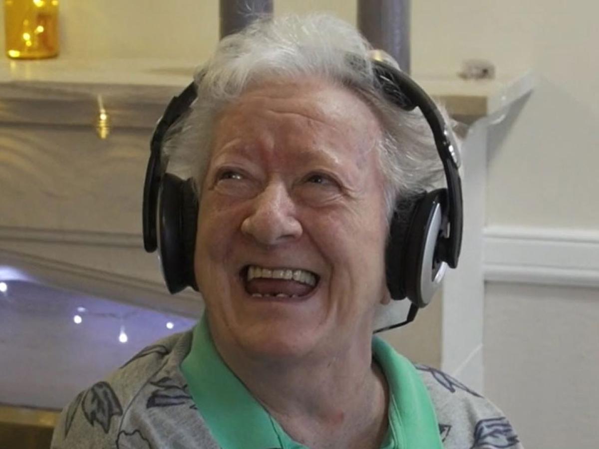 An elderly woman wearing headphone looks up and to the side with a big smile.