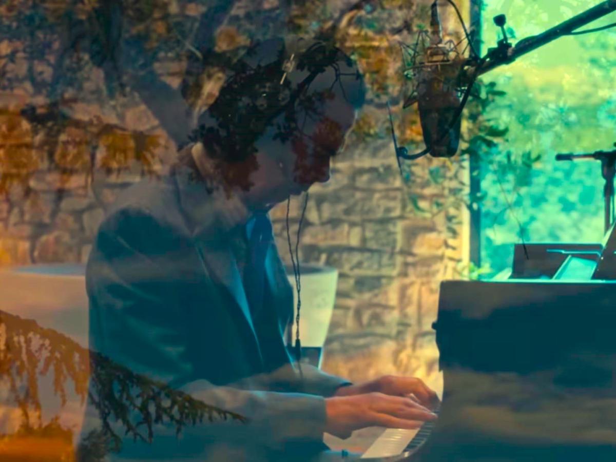 A pianist raises his arms while sitting at a grand piano amid recording equipment.