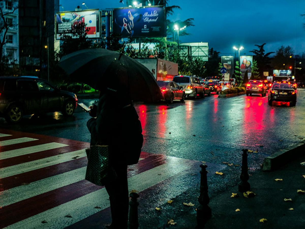 On a rainy night a pedestrian, holding a brolly, waits to cross a road.