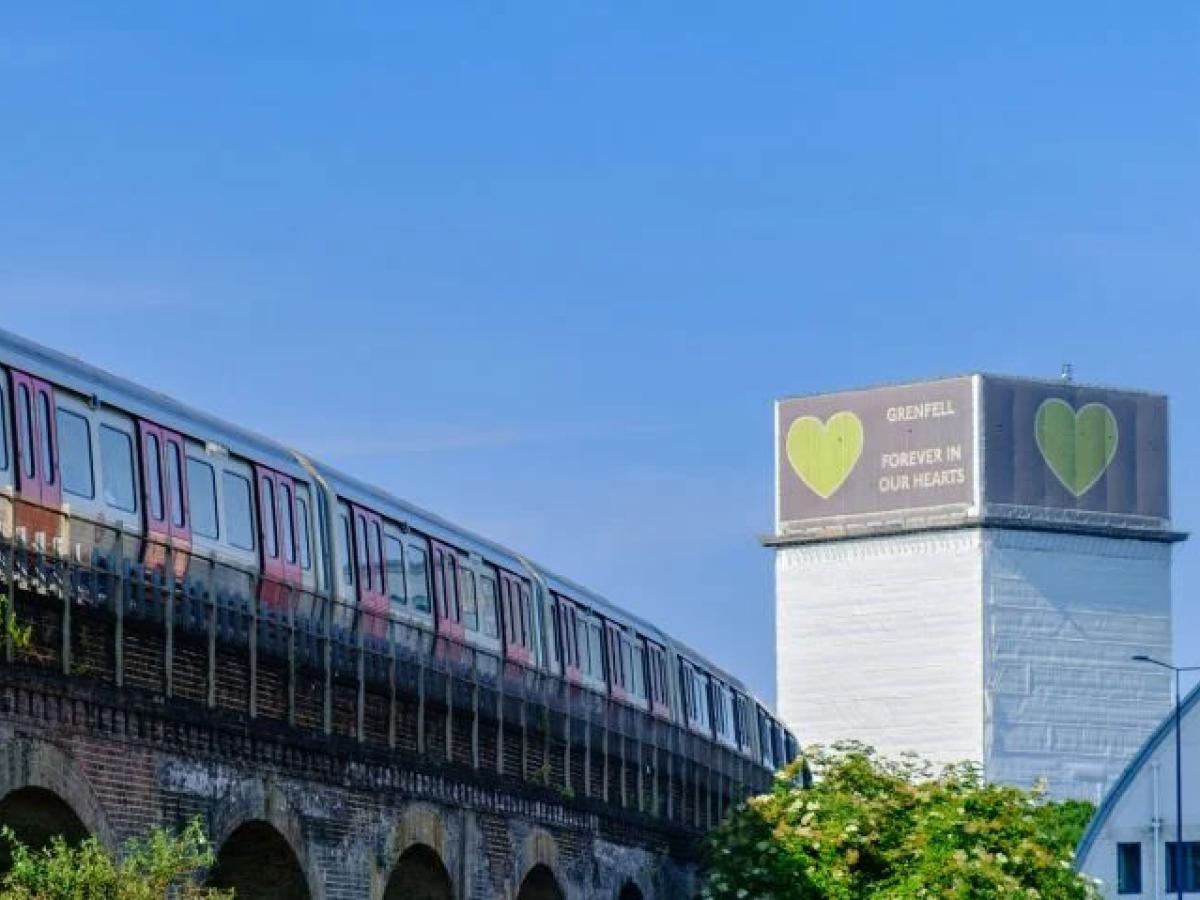 A tube train runs on a raised track, in the distance is a tower block wrapped in white material with a green heart on it.