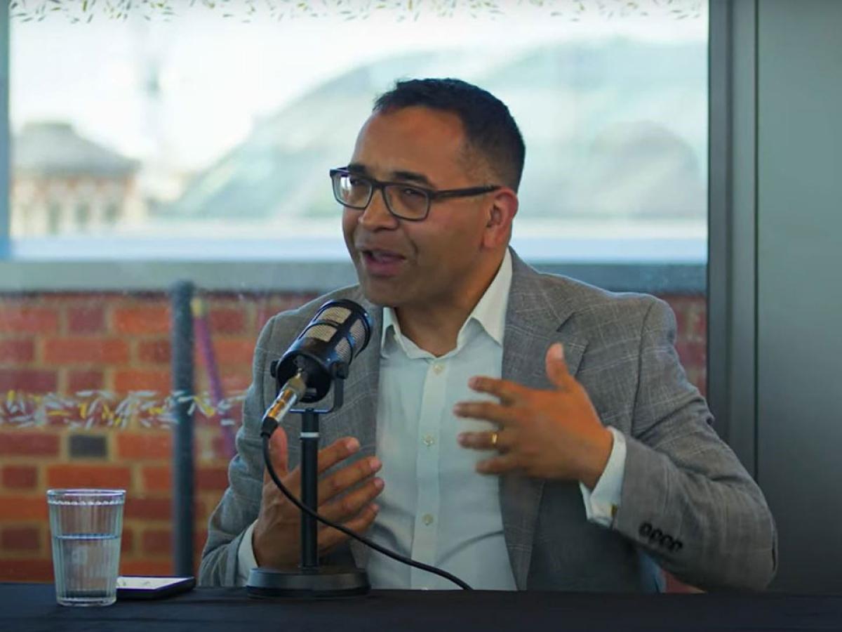 A man sitting at a table with a mic on it, raises his hands in gesture as he speaks. 