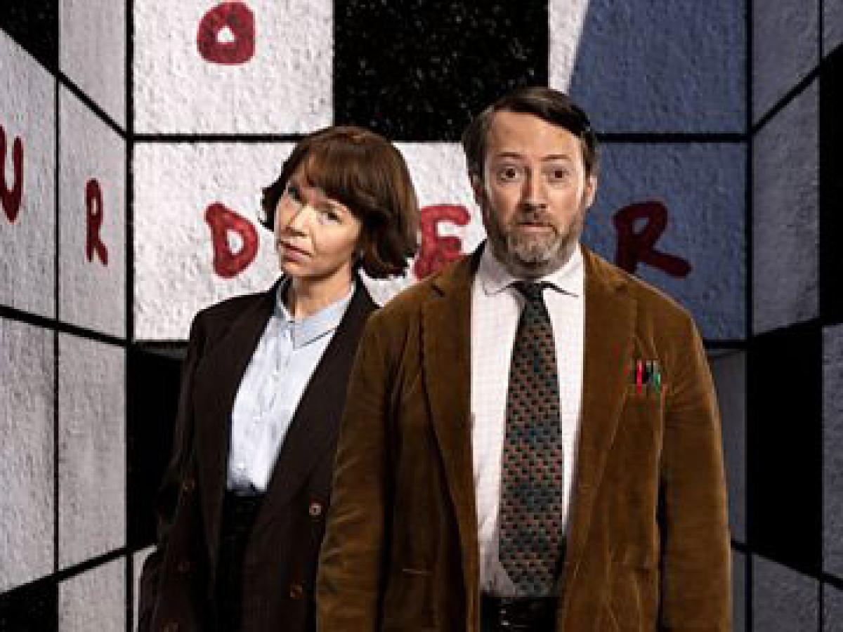 Two TV characters, a man and a woman, stand in front of a crossword cover walls of a room. 