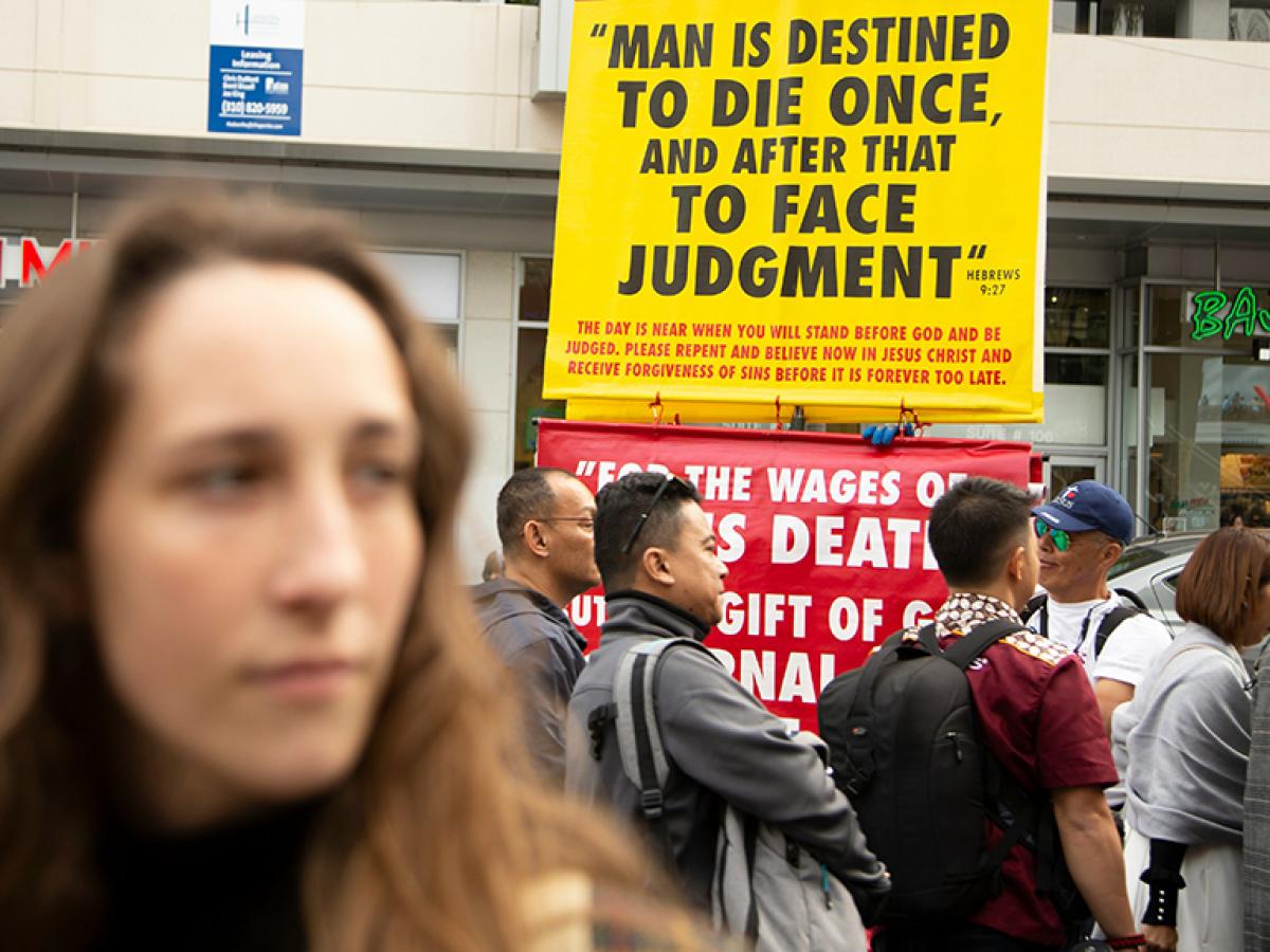 Behind a passer by a street peacher holds up a large yellow sign with a message on it.