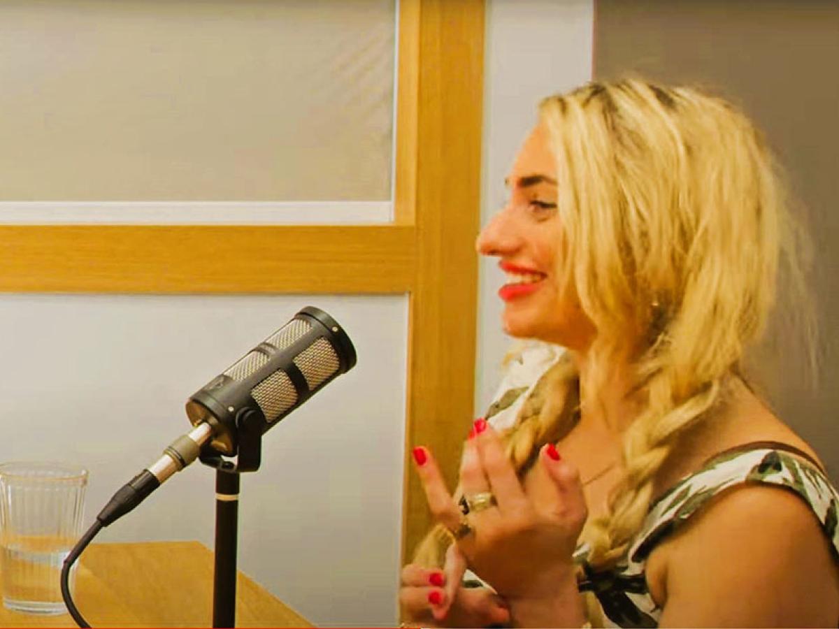  In profile a woman speaks into a microphone and holds her hands up from a desk.