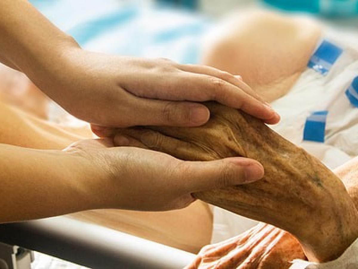 A younger hand holds a wrinkled older hand of someone in a bed.