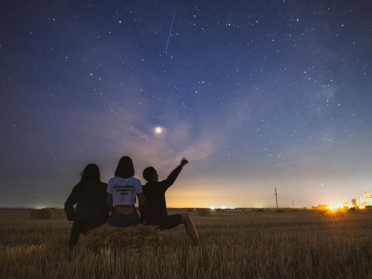 At dusk, three people sit on a field edge and look at the stars emerging.