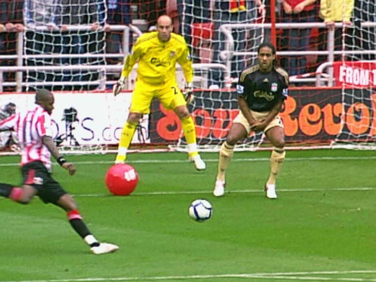 A striker is about kick a football towards a goal, a red beach ball sits between him and the goalkeeper.