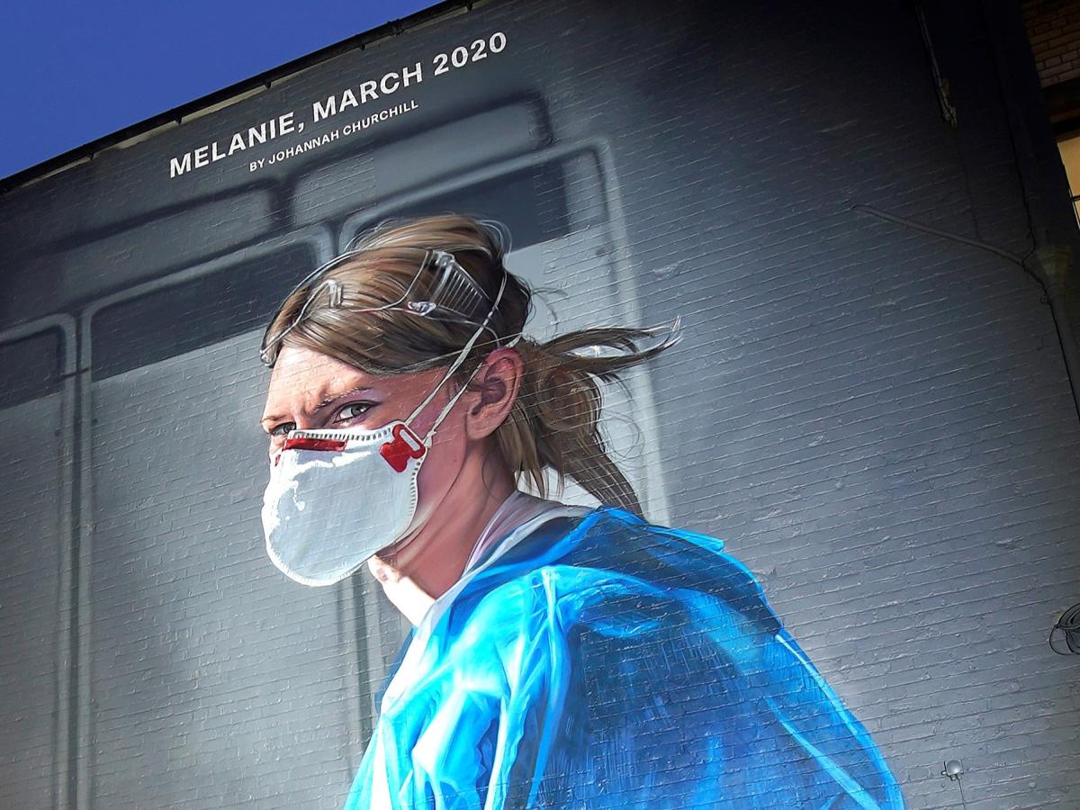 A gable end mural depicts a nurse in scrubs and a mask turning and looking towards the viewer.