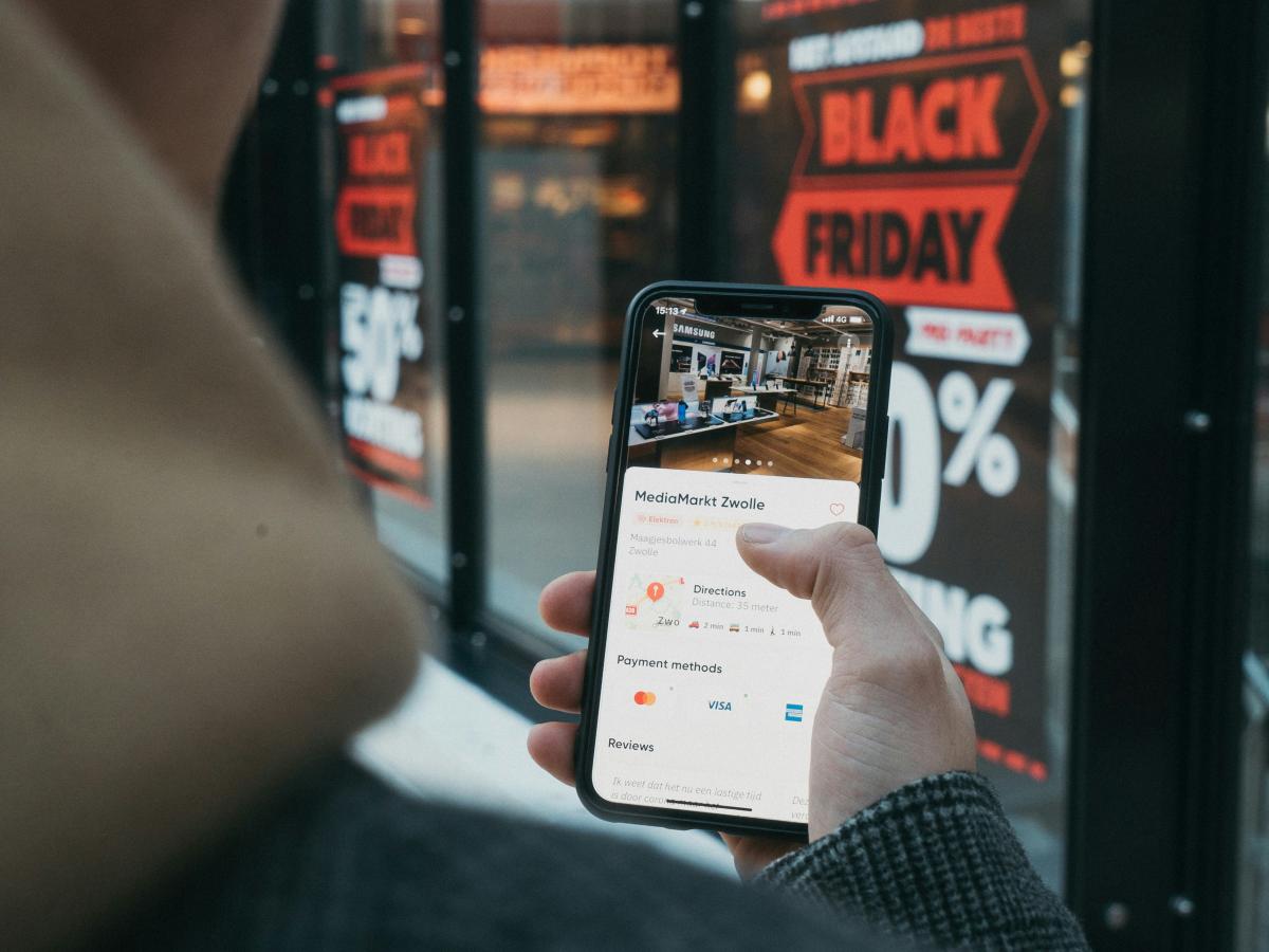 A phone screen is held up, showing a shopping page, behind a window displays a Black Friday Sale banner.