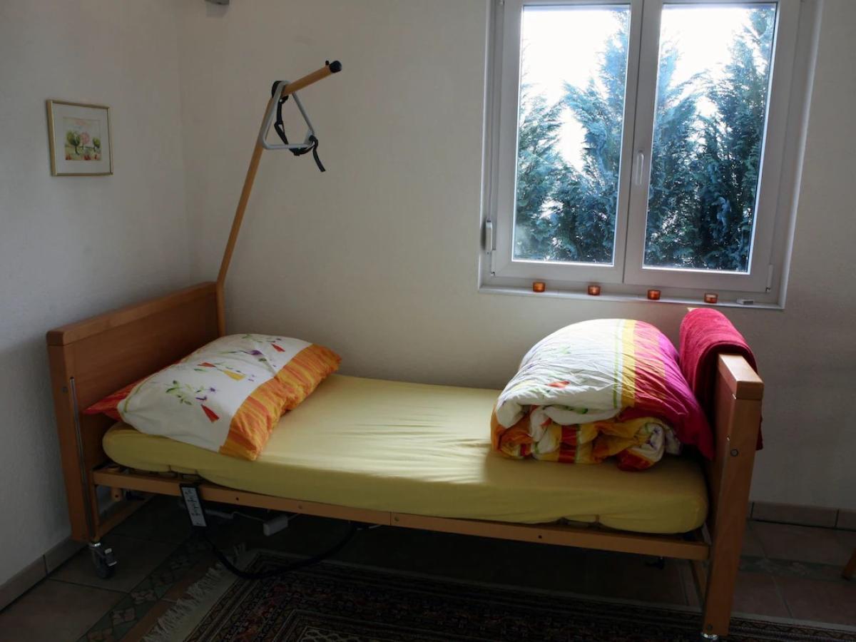 A single bed, wiith an unmade colourful duvet stands in the corner of a room. A hoist reaches over it from the corner.