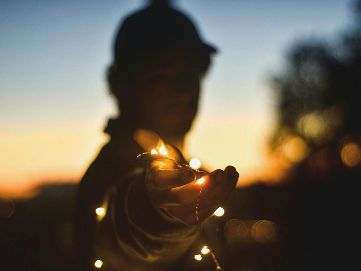 A backlit person at twilight holds a hand out towards the camera, holding some fairly lights