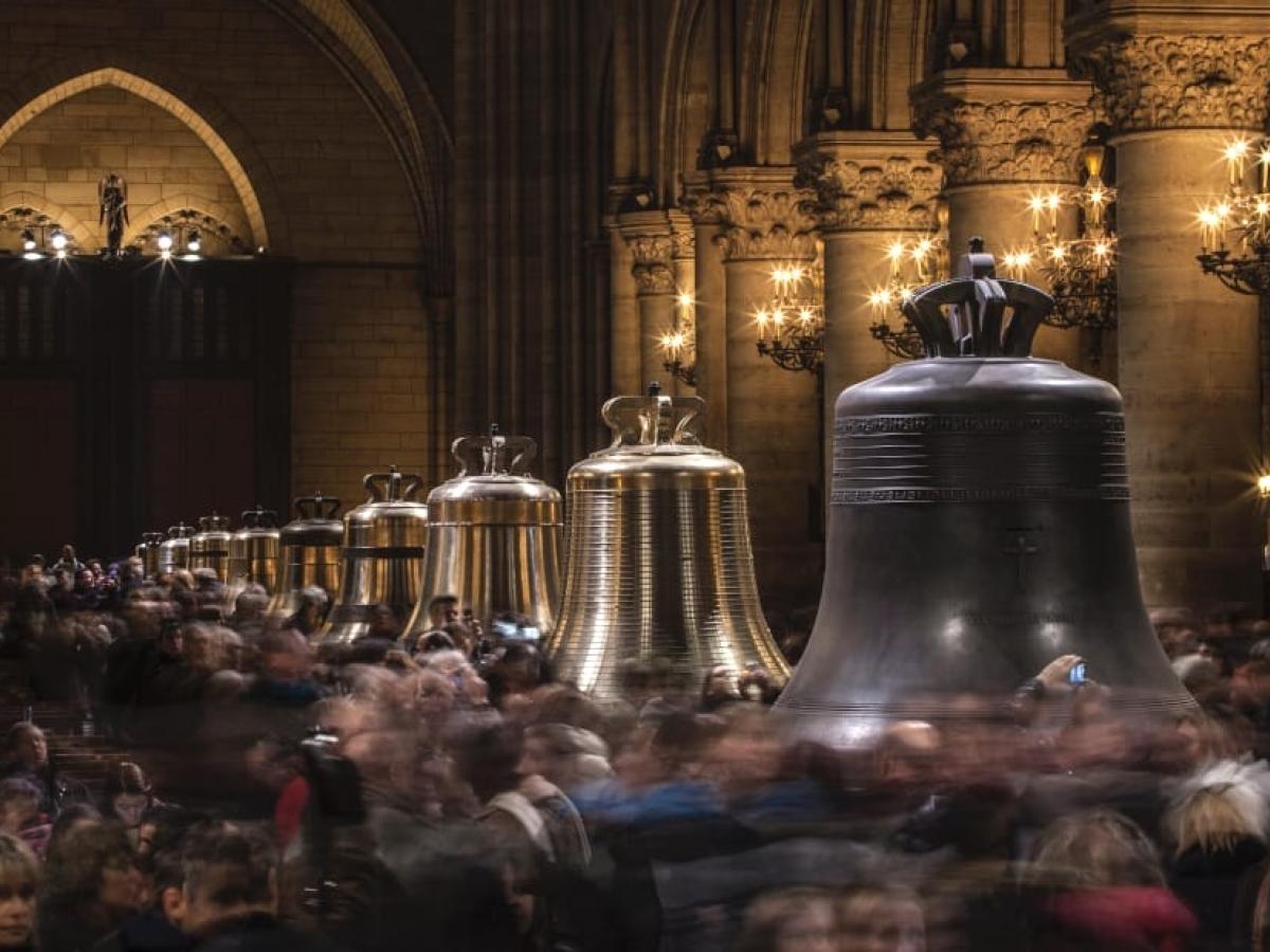 Restored church bells lined up in a cathedral, as crowds mill around them.