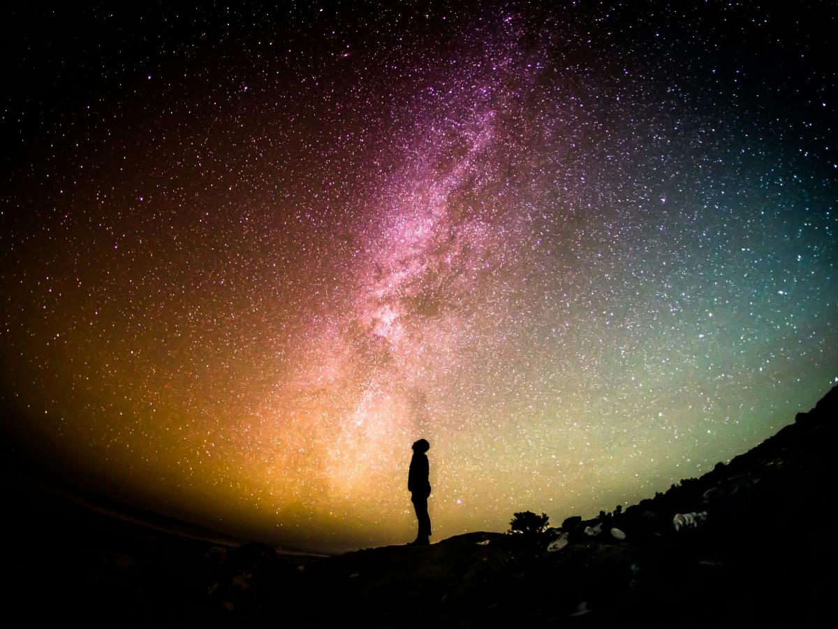 A fish eye lens view of a person standing silhouetted, looking up to a colourful night sky with the Milky Way across it