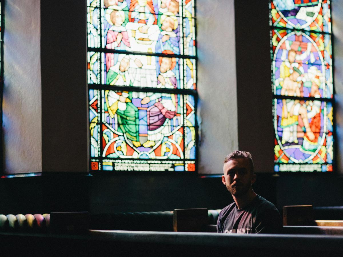 A man sits in a church pew below a colourful stained glass window, looking pensive.