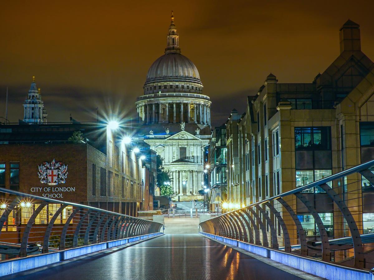 A view from a floodlight footbridge towards a gap between office buildings which reveal a cathedral and its illuminated dome.