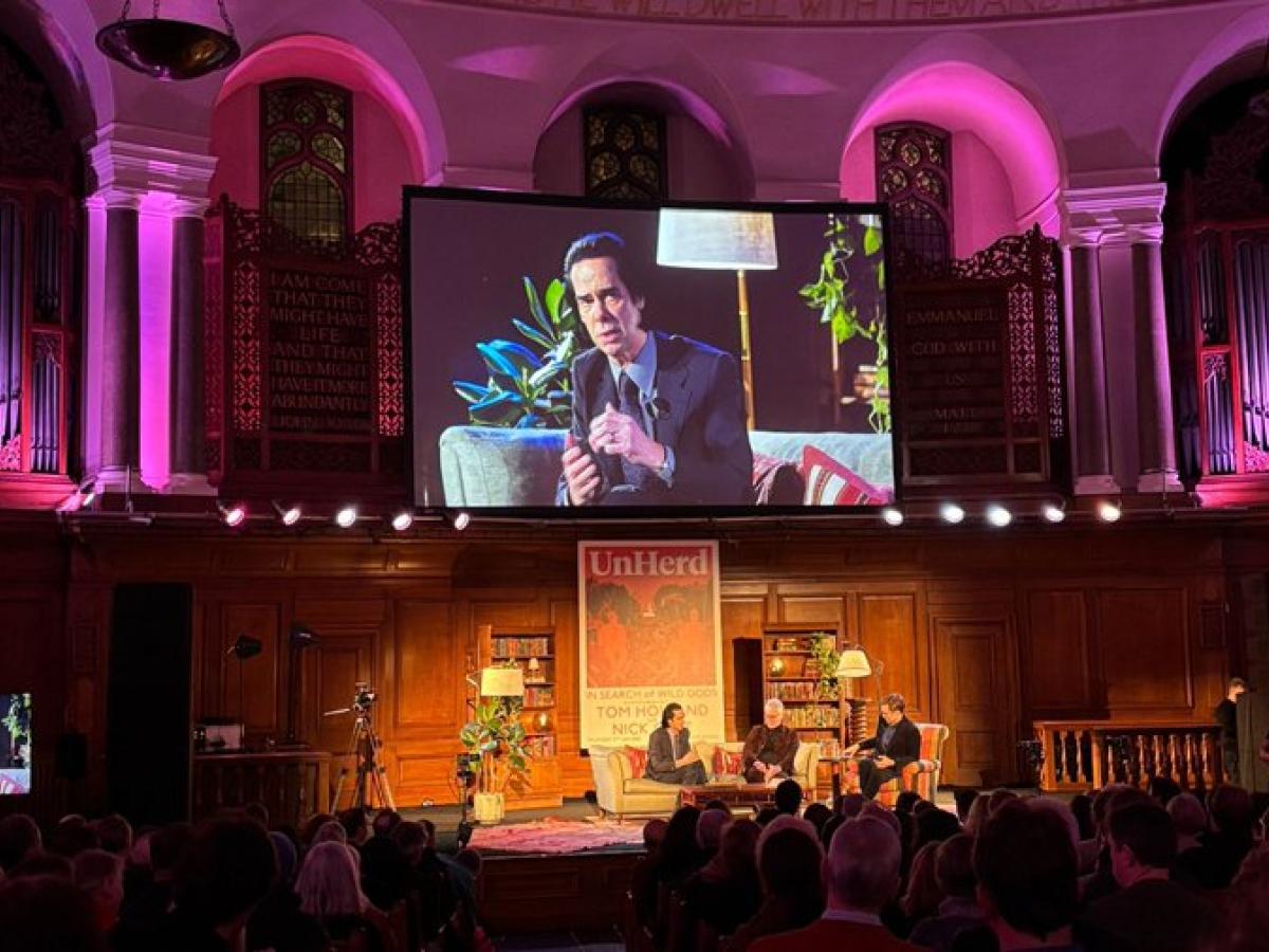 A view of an in-conversation on a stage, with a video screen above showing a close up.