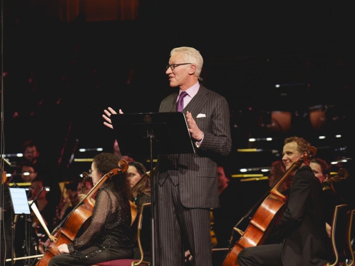 A man in a suit stands in front of a orchestra, by a lectern, gesturing while talking.