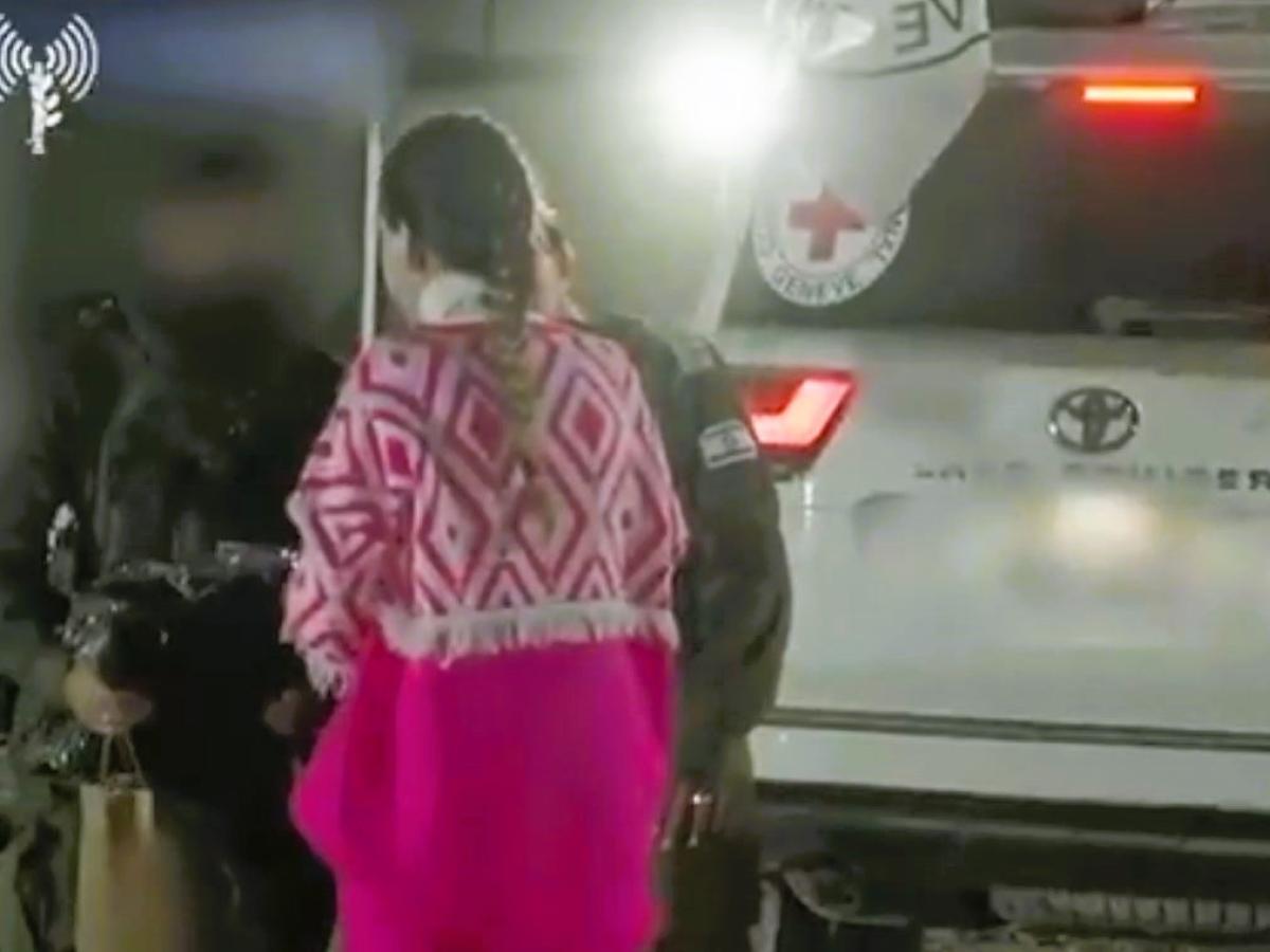 At a night time hostage exchange, a hostage wears a pink top and is guide toward the side of a white vehicle bearing a small red cross.