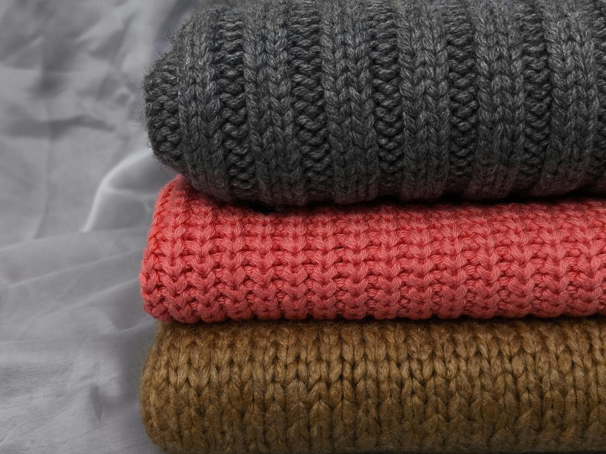 A pile of folded knitted jumpers sit on a grey back ground, one is also grey, the others are red and brown.