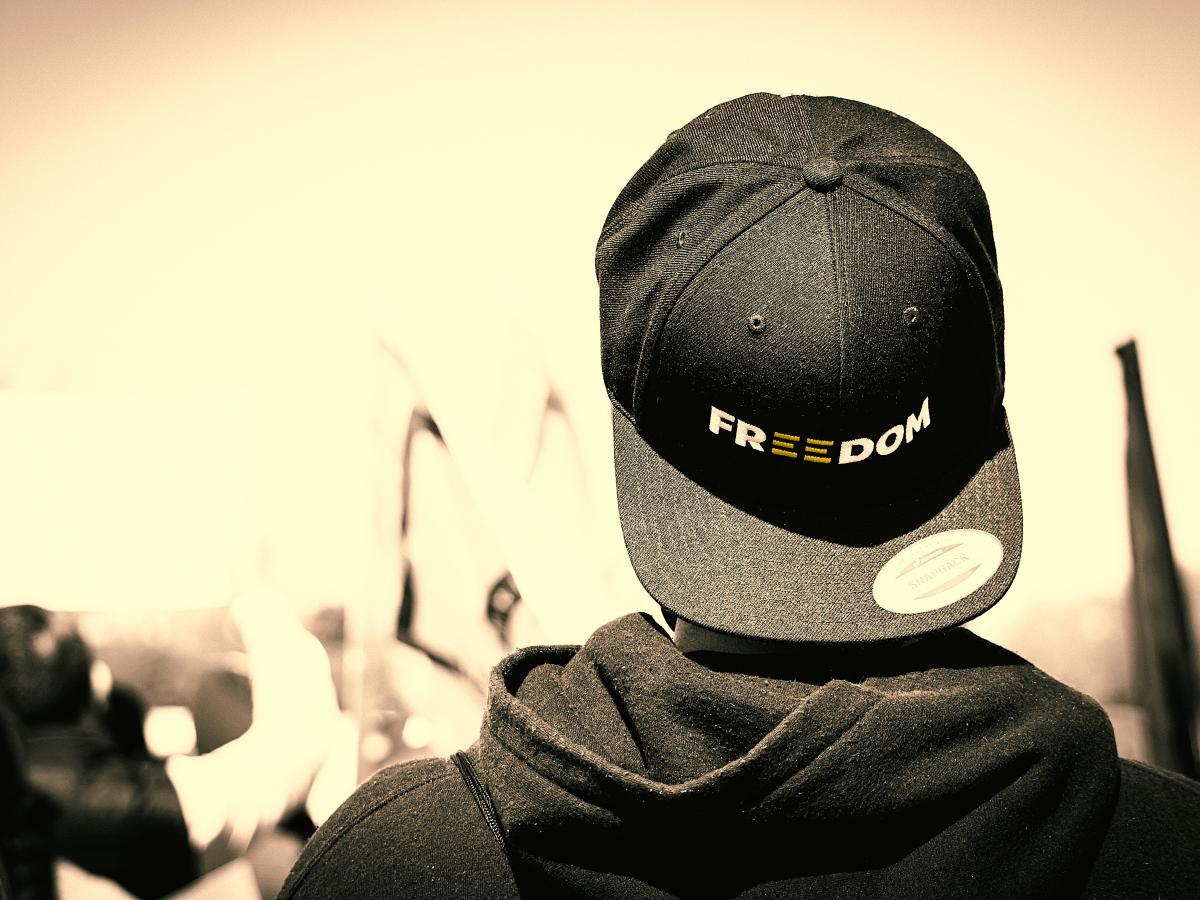 A protester stands with back to the camera, his baseball hat is turned backwards, it reads 'freedom'.