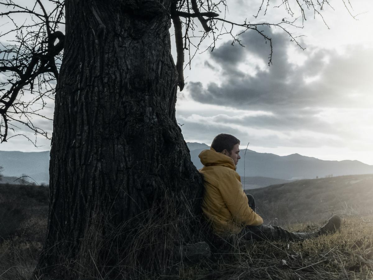 A wrapped-up man sits and leans against a bare tree, as dark clouds give way to sun.