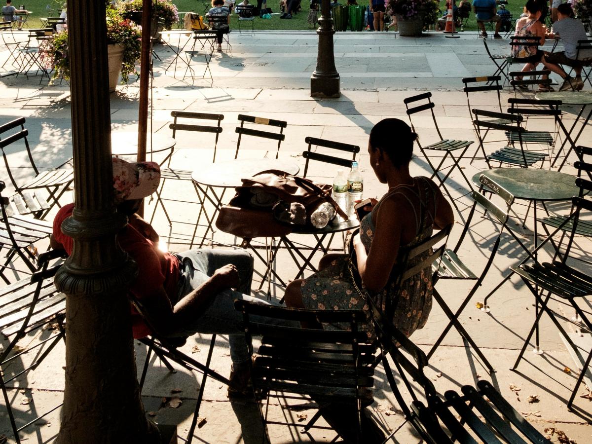 Two people. sitting at a street cafe amid empty tables and chairs, are silhoutted.