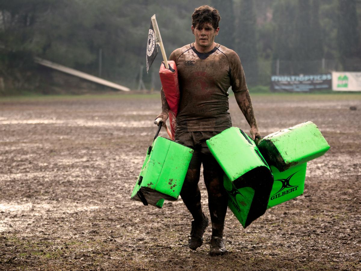 A muddy rugby player carries equipment off a pitch.