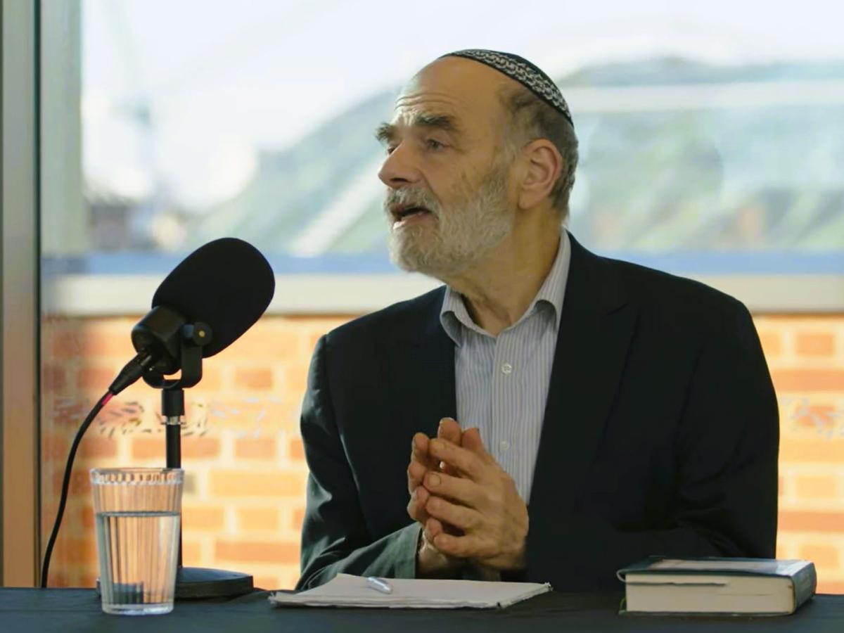 A podcast guest holds his hands together in front of himself as he raise his head in speech.