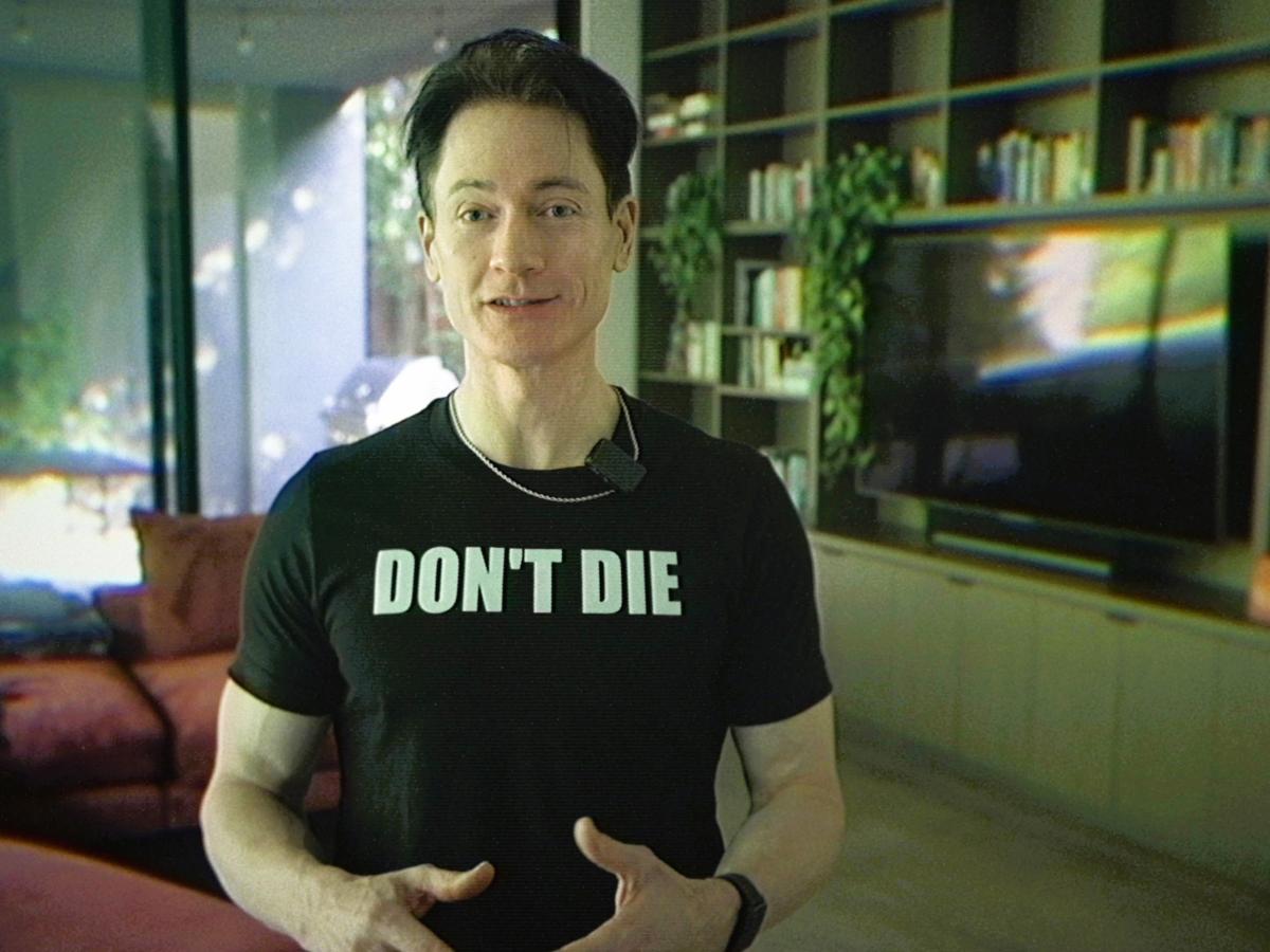A man stands in his home wearing a black t-shirt that reads 'DON'T DIE'.