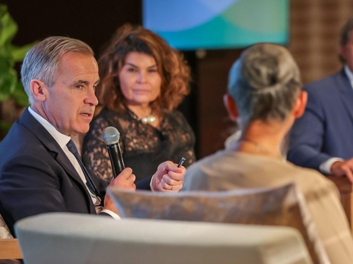 Mark Carney sits between two other speakers, holding a mic.