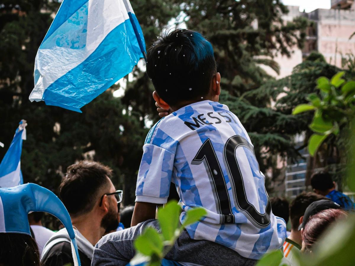 A child on the shoulders of a parent wears a light blue and white stripped football top, waves the Argentinian flag