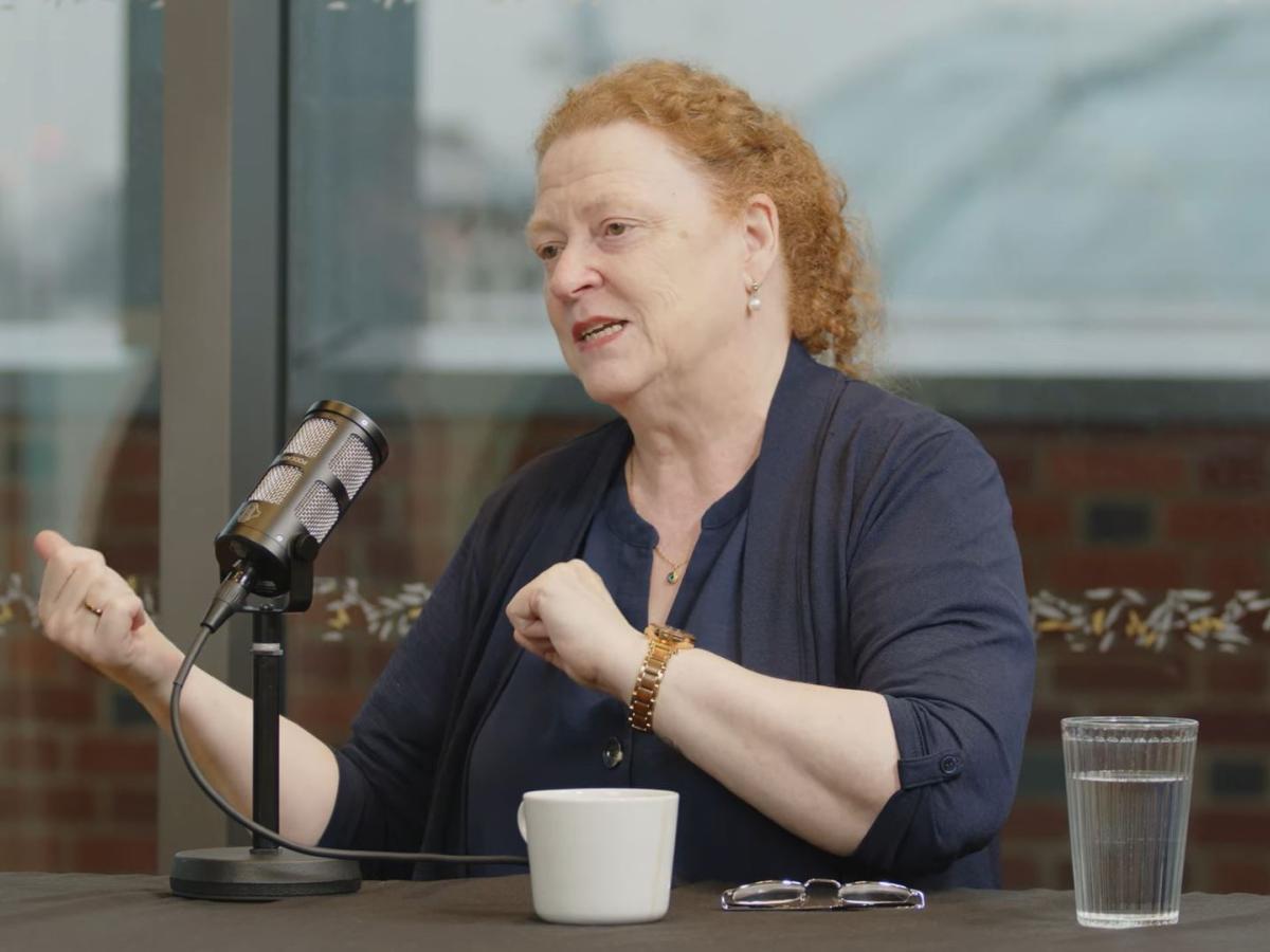 A woman guest on a podcast sits at a table with a mic in front, gesturing to the side with both hands.