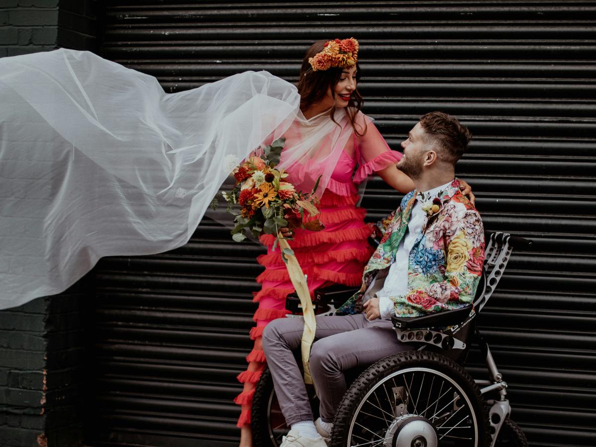 A bride dressed colourfully stands next to her groom, dressed similarly, as he sits in a wheelchair.