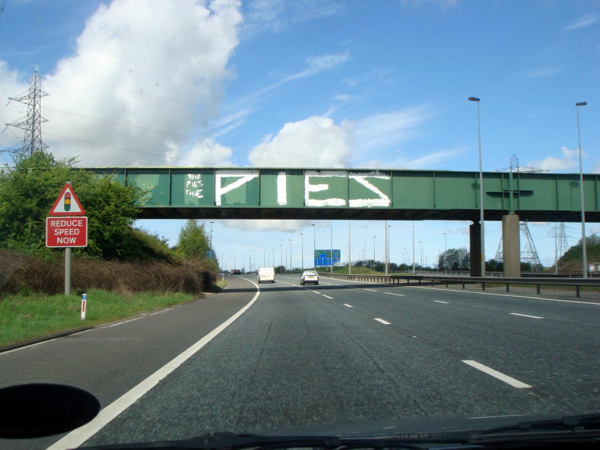 A green bridge spans a motorway, on its side is graffiti that reads 'PIES'