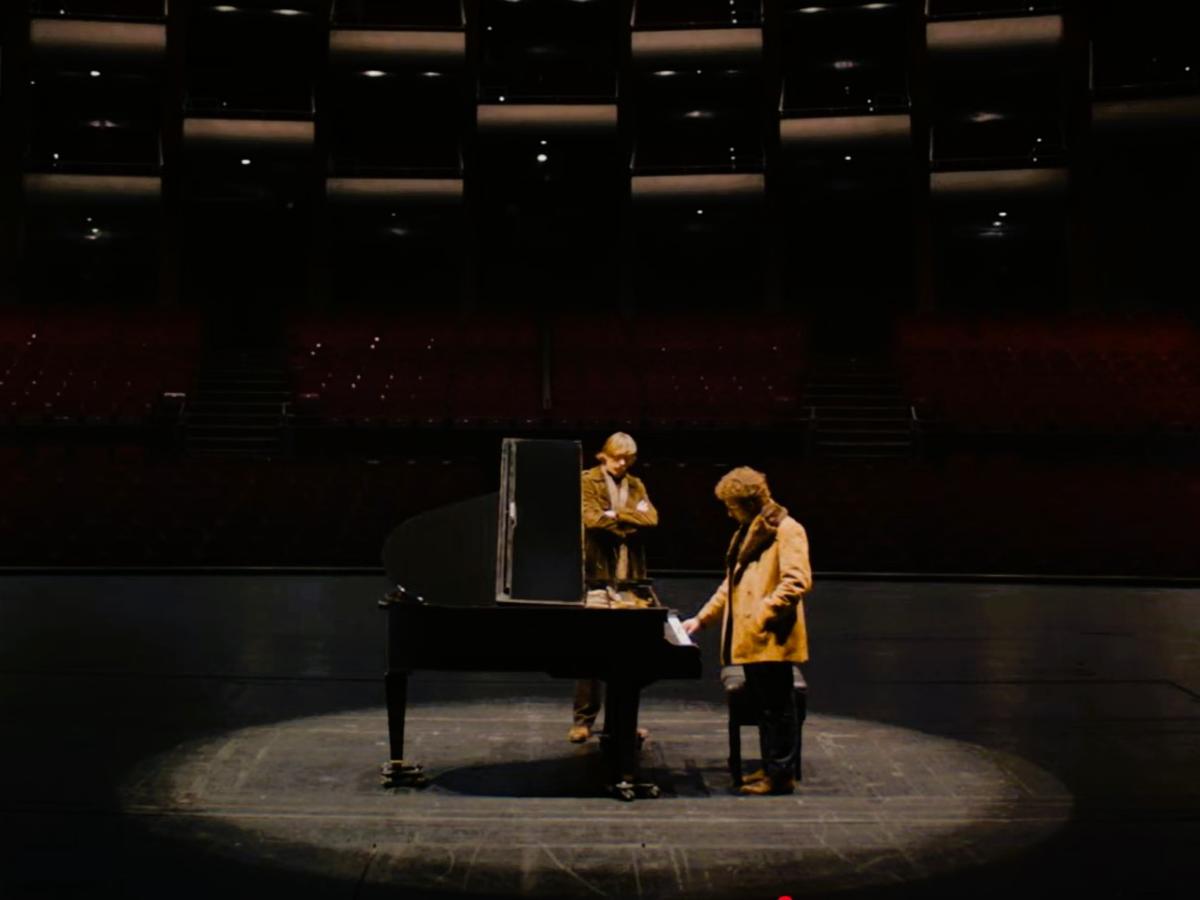 Two people sit and stand next to a grand piano on a stage.