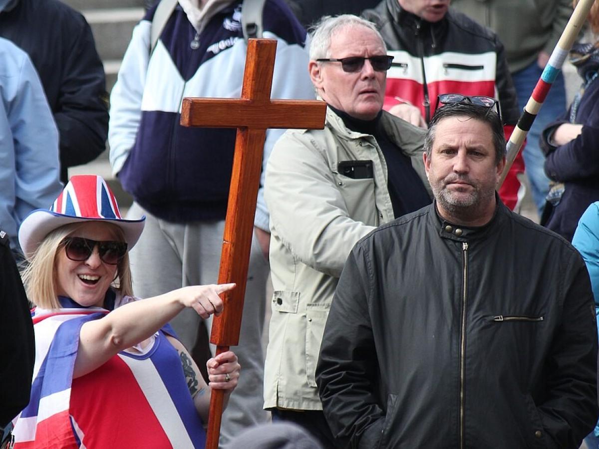A protester wearing a Union Jack flag and hat and holding a cross, points while a man looks on.