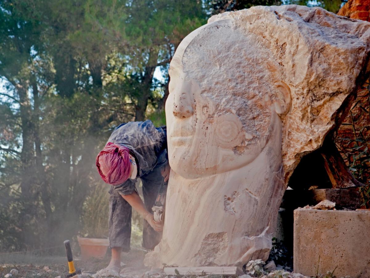 A sculptor works on a carved face emerging from a stone larger than her