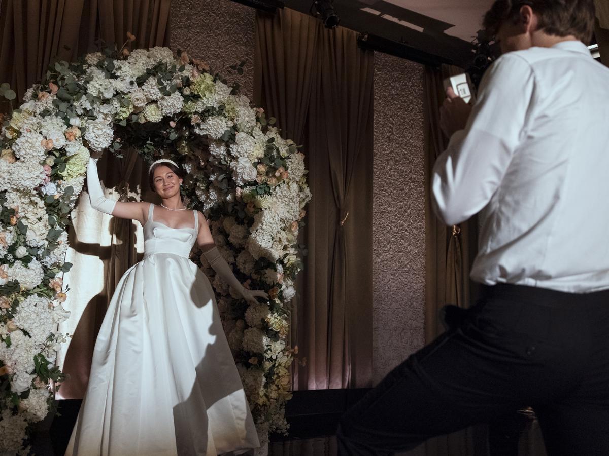 A woman in a white dress stand in front of a large floral arrangement.