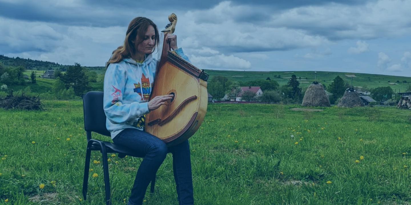 A woman sits on a chair in a field holding a large stringed musical instrument.