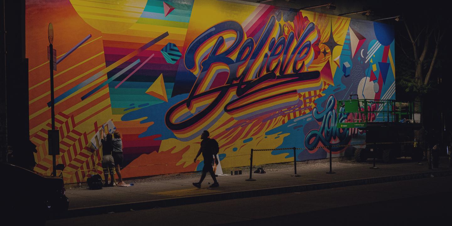 A person walks past a multi-coloured wall of graffiti with the word 'believe' in the centre of it.