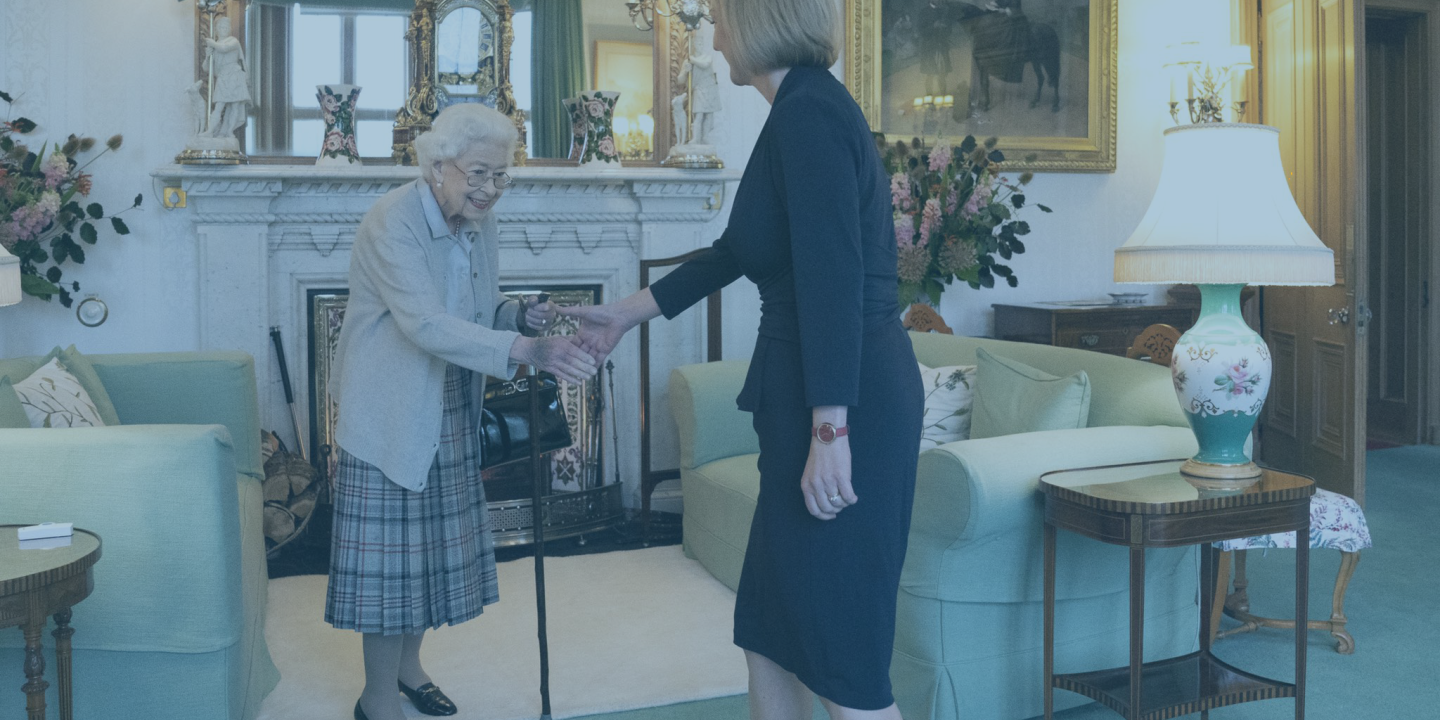 A frail old lady, the late Queen, rises from a sofa to shake hands with an approaching woman.